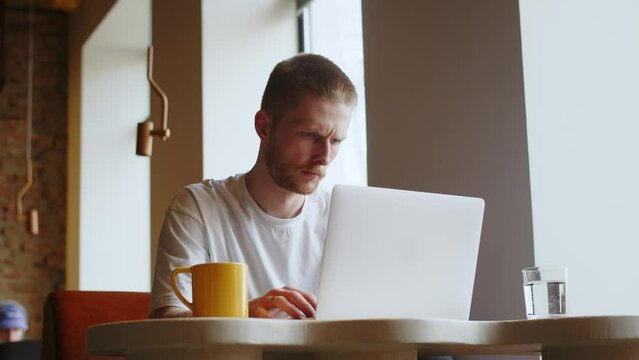 A frowning man is typing something on the keyboard of his laptop. A man in a white T-shirt works remotely at his computer, sitting at a table in a cafe.