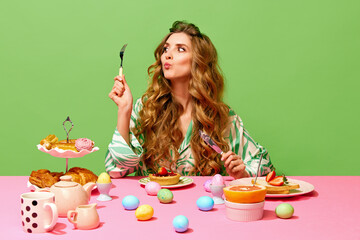 Food pop art of beautiful girl with long hair sitting at table holding cutlery and eating over green background. Colorful eggs on table