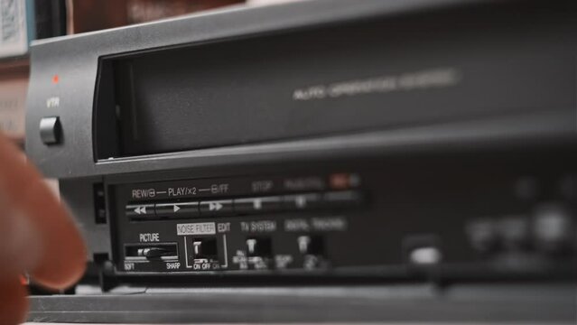 Male hand inserts VHS cassette into a VCR Video recorder. Black vintage videotape cassette recorder on desk with many video cassettes. Inserting retro VHS tape into vintage player. Home video archive