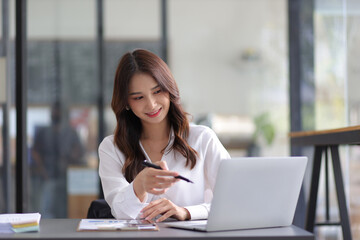 Young beautiful asian businesswoman in office working with laptop and financial documents on desk.