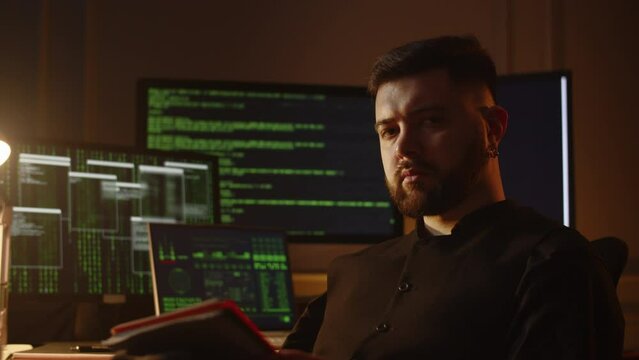 View of a young dark-haired man with a serious look. A male programmer sits in his room with a notebook in his hands against the background of a monitor with running lines of program code.