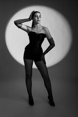 A sexy young woman in a black corset, gloves and underwear poses against the background of a light circle. Black and white photo of a sexy young woman in full growth.