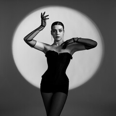 A sexy young woman in a black corset, gloves and underwear poses against the background of a light circle. Black and white photo of a sexy young woman.