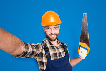 Portrait of handsome cheerful master in overall, shirt, hardhat, uniform shooting selfie on front camera, showing saw in arm, having leisure fun, isolated on grey background