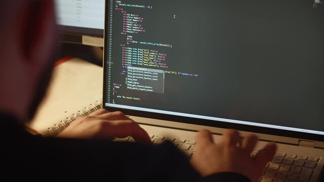 An experienced web developer writes the code for a website. A male programmer is working on his laptop in the office. View from behind.