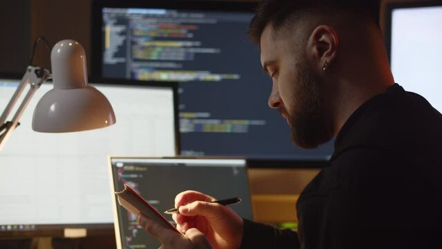 A young man is learning programming on his own while sitting at home. A man sits in front of the monitors on which the program code is visible and makes notes in his notebook.