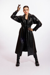 A sexy young woman in a long leather raincoat and bright makeup poses on a white background.