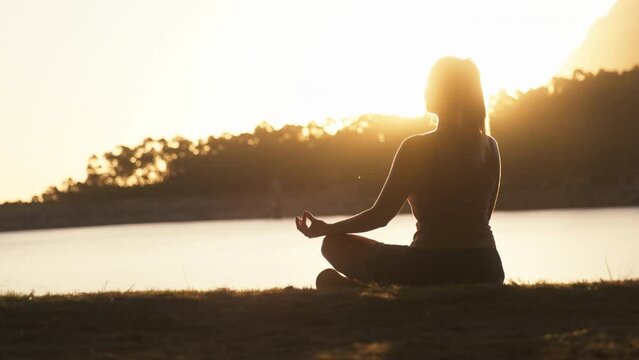Woman relaxing sitting by beautiful lake and mountains at sunset doing yoga - shot in slow motion