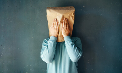 Man with a paper bag on head covering eyes with both hands over grey background