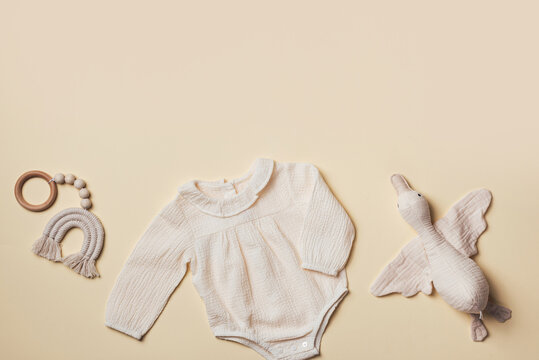 Mockup of cream infant bodysuit made of organic cotton with eco friendly baby accessories, knitted rainbow, soft duck on beige backgroundd. Gift for newborn baby. Top view. Flat lay. Copy space