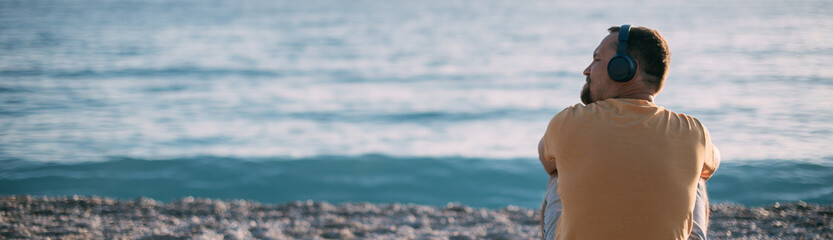 Portrait of a young man sitting on the seashore with headphones at sunset.