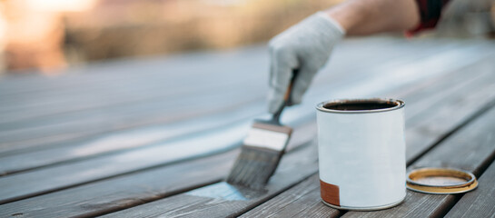 Close-up, an open jar and a man's hand in a work glove with a painting brush paints boards outdoors.