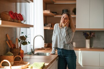 Standing and talking by phone. Beautiful young woman is on the kitchen