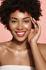 Vertical shot of young curly woman, beauty model with glowing facial skin, using skincare product, washing her face with cleanser, smiling at camera, pink background