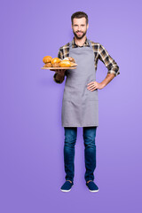 Full size body portrait of positive attractive baker in jeans, shoes, shirt, apron with stubble having tray with bakery products, looking at camera, isolated on grey background