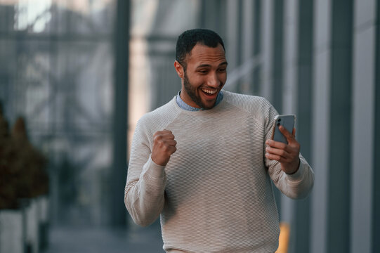 Happy guy holding smartphone and celebrating success. Handsome black man is outdoors near the business building - Powered by Adobe