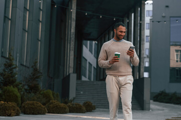 With cup of coffee and smartphone. Handsome black man is outdoors near the business building