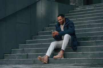 Sitting on the stairs with smartphone in hands. Handsome black man is outdoors near the business building