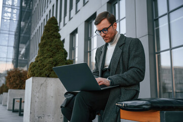 Sitting on the bench with laptop. Handsome man in formal clothes is outdoor near the business building