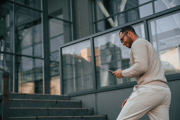 Against modern business building. With smarpthone. Handsome black man is outdoors