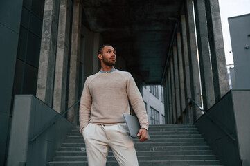 Front view. Standing with laptop in hands. Handsome black man is outdoors near the business building - Powered by Adobe