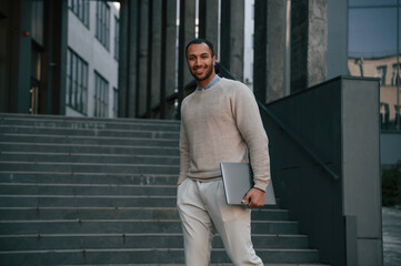 Fototapeta premium With laptop in hands. Handsome black man is outdoors near the business building