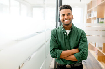 Handsome indian male office employee standing with his arms crossed, looking at the camera and laughs. Portrait of cheerful young man wearing smart casual shirt in modern coworking space - obrazy, fototapety, plakaty