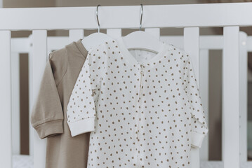 Beige baby bodysuit with hangers on bed, beige background. Cute children's clothes in nursery room....