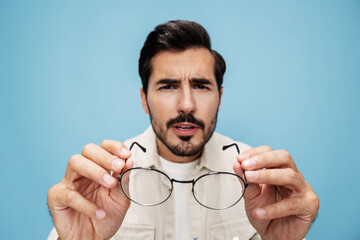 Close-up portrait of a brunette man looking through glasses that he holds in his hands, eye problems, glasses for vision farsightedness and nearsightedness, on a blue background, copy space 