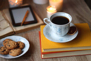 Cup of tea or coffee, plate of cookies, books, e-reader, pencil and lit candles on the table. Selective focus.
