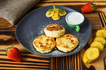 Syrniki or cheese pancakes with cream sauce and bananas on a plate on wooden table close up