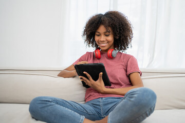 Happy millennial African teen girl checking social media holding tablet at home app playing game, shopping online, ordering delivery relax on sofa.
