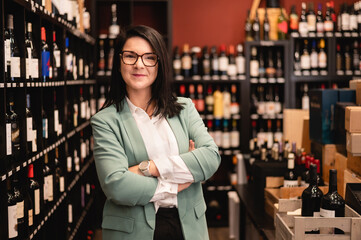 Smiling adult female manager wearing green jacket and white shirt standing in liquor store with...