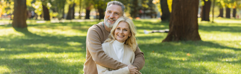 overjoyed middle aged man with grey beard hugging charming blonde wife in park, banner.