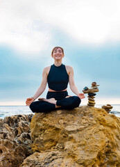 young female is sitting in yoga lotus pose on the stone near the ocean