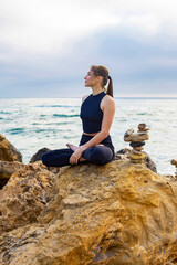 young female is sitting in yoga lotus pose on the stone near the ocean
