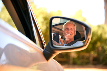 Fototapeta na wymiar Senior Male Passenger With Mobile Phone Reflected In Wing Mirror Of Car Enjoying Day Trip Out