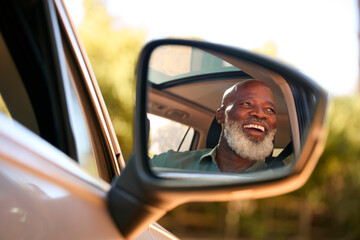 Smiling Senior Male Driver Reflected In Wing Mirror Of Car Enjoying Day Trip Out Driving - obrazy, fototapety, plakaty