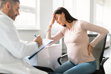 Sick pregnant lady feeling unwell, touching head and back during meeting with her doctor in clinic