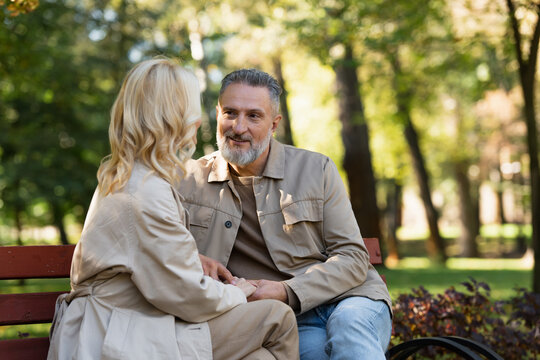 Smiling mature man holding hand of blonde wife while sitting on bench in park.