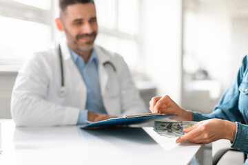 Female patient putting money under doctor's clipboard, sitting at table in clinic, man looking at it and smiling
