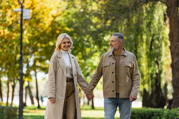 Happy mature man holding hand of blonde wife in trench coat in park.