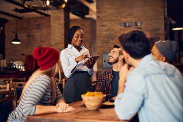 Happy black waitress using touchpad while taking order from her customers in bar.