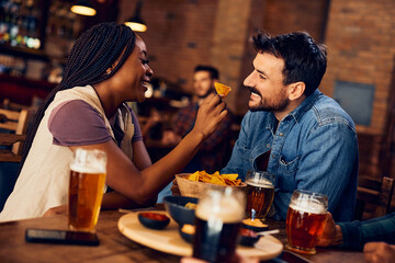 Happy black woman has fun while feeding her boyfriend with nacho chips in pub.
