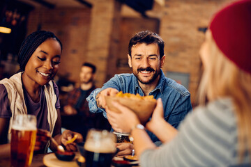 Happy man and his friends eat nacho chips and drink beer while gathering in bar.