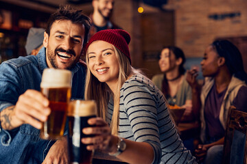 Young couple has fun while toasting with beer in bar and looking at camera.