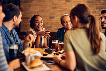 Happy black woman talks to her friends while drinking beer in pub.