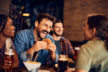 Happy man eats burger while talking to his friends in pub. - obrazy, fototapety, plakaty