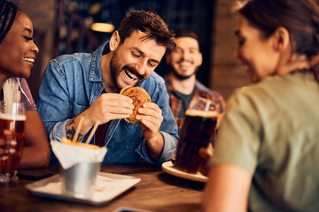 Happy man eats burger while gathering with friends in bar.