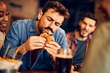 Hungry man eats burger while gathering with friends in pub.